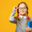 Schulmädchen (etwa 7 Jahre alt) vor einem gelben Hintergrund. Sie trägt eine Brille, die sie mit einer Hand festhält, hat zwei blonde Zöpfe mit rten Schleifen und hält ein blaues Buch in der anderen Hand. Sie lächelt in die Kamera.
