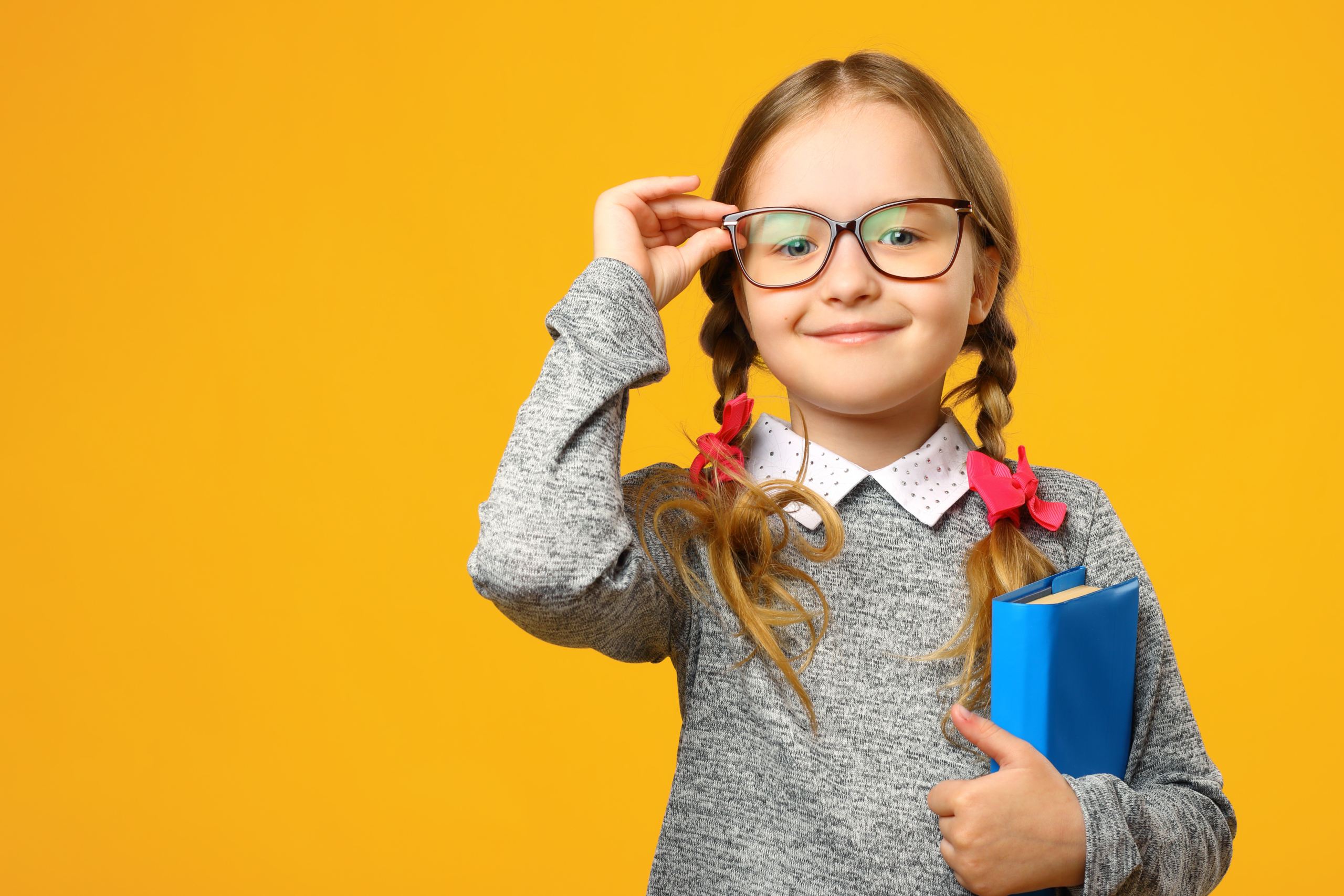 Schulmädchen (etwa 7 Jahre alt) vor einem gelben Hintergrund. Sie trägt eine Brille, die sie mit einer Hand festhält, hat zwei blonde Zöpfe mit rten Schleifen und hält ein blaues Buch in der anderen Hand. Sie lächelt in die Kamera.