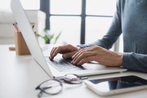 Schreibtisch mit einem Laptop im FOkus, seitlich. Es sind nur tippende Hände auf der Tastatur zu sehen. Neben dem Laptop liegen eine Brille und ein Smartphone