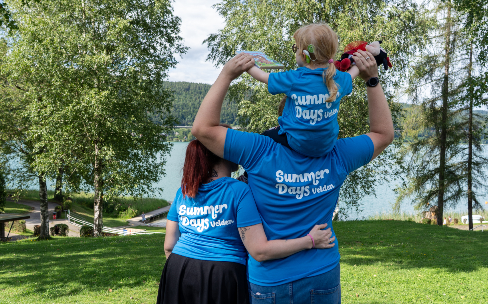 Eine Familie steht mit dem Rücken zur Kamera. Mutter, Vater, Tochter. Die Tochter sitzt auf den Schultern des Vaters. Alle tragen ein blaues Shirt, auf dem Summer Days Velden steht.