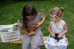 zwei Mädchen sitzen auf einer Bank, beide haben ein Kaninchen bzw. ein Meerschweinchen im Schoß. Das rechte Mädchen trägt ein Cochlea-Implantat und schaut zum Mädchen links rüber.
