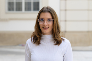 POrträtfoto von Stefanie Muck. Eine junge Frau, Ende 20, weiß mit hellbraunem Haar und Brille. Sie trägt einen weißen ROllkragenpulli und lächelt freundlich in die Kamera.