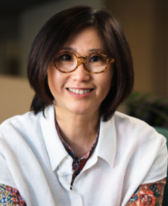 Porträtfoto von Yin Ge. Eine asiatisch aussehende Frau in den 40ern lächelt freundlich in die Kamera. Sie trägt eine weiße Bluse, eine runde Hornbrille und hat einen dunkelbraunen Bob.